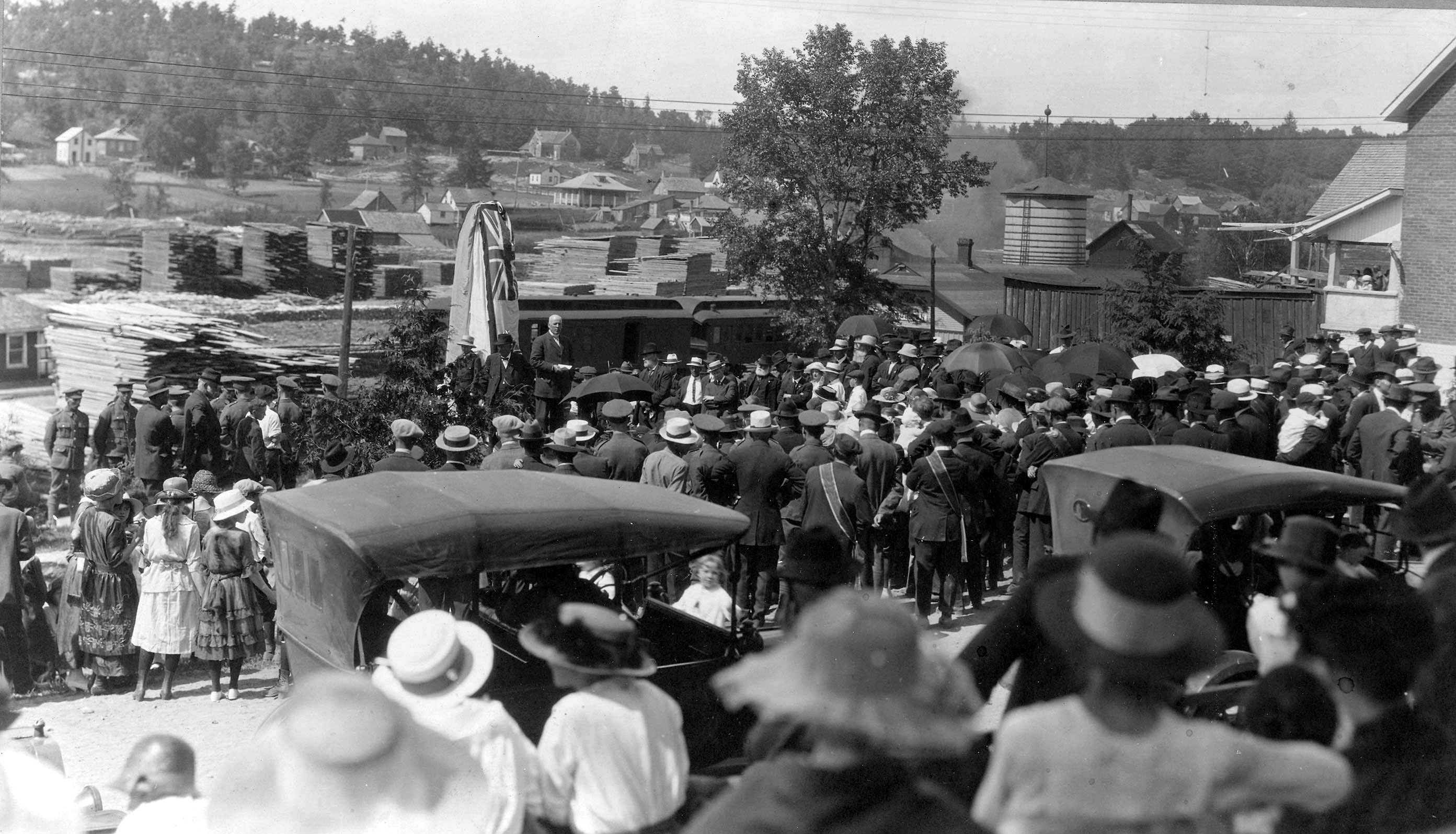 Crowd of people at the unveiling of the Cenotaph. There are piles of lumber in the background and a few cards in the foreground.