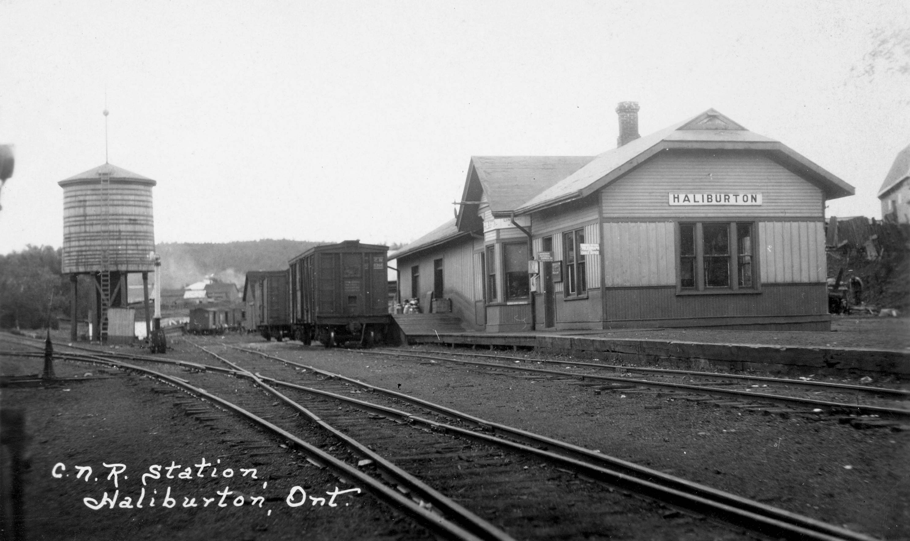 Railway tracks, with box cards parked in front of the railway station. To the left of the picture there is a water tower with a ladder up the side.