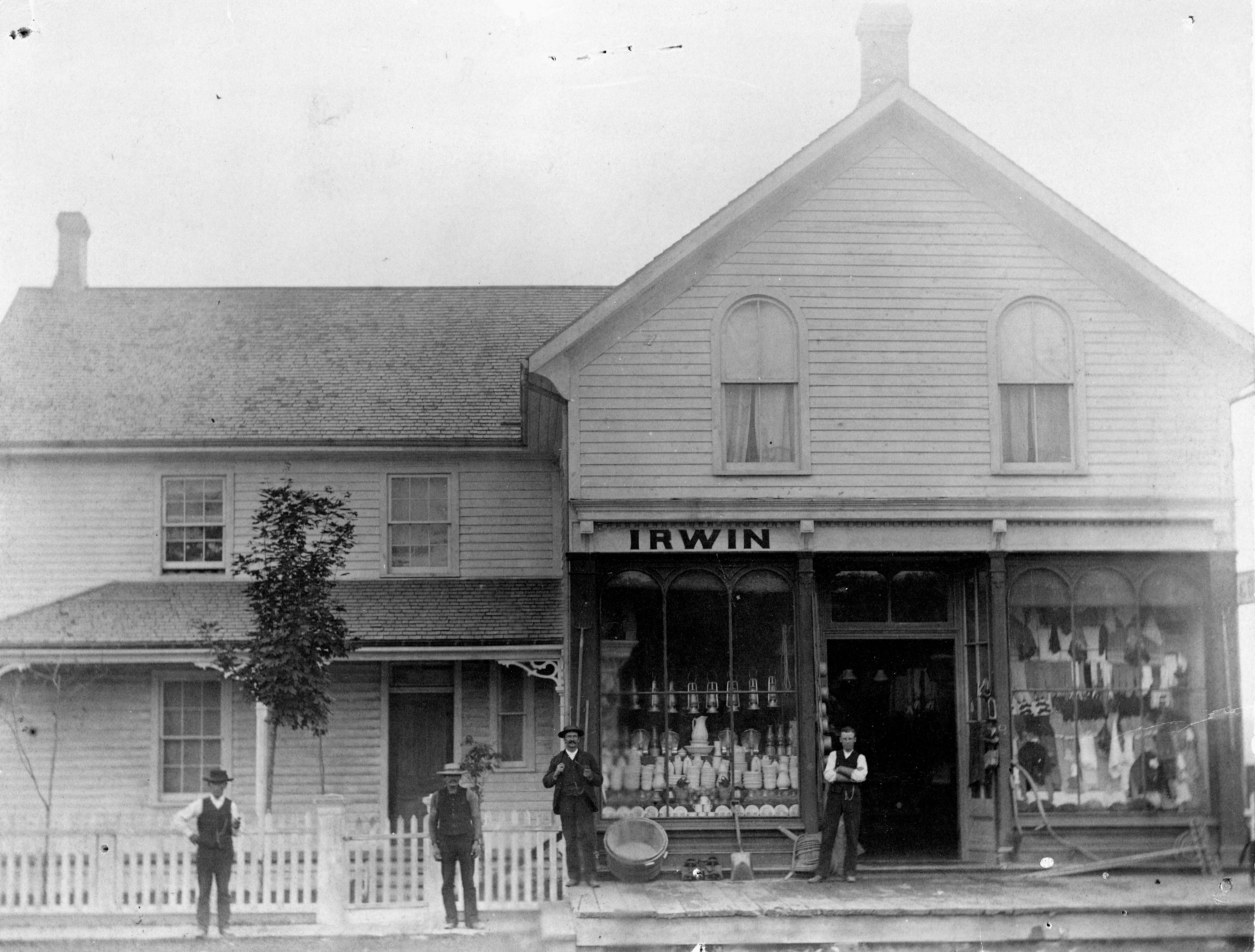 Black and white photo of s store with two large glass store front windows, a white picket fence and four men dressed in dark pants, white shirts, top hats and vests.