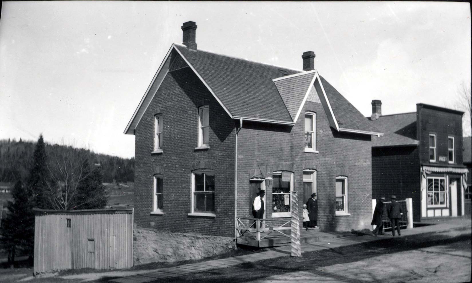 Two story red brick home with two entrance doors. In front of the one door, there is a wooden barbershop pole and a man. In front of the other door there is a woman and child.