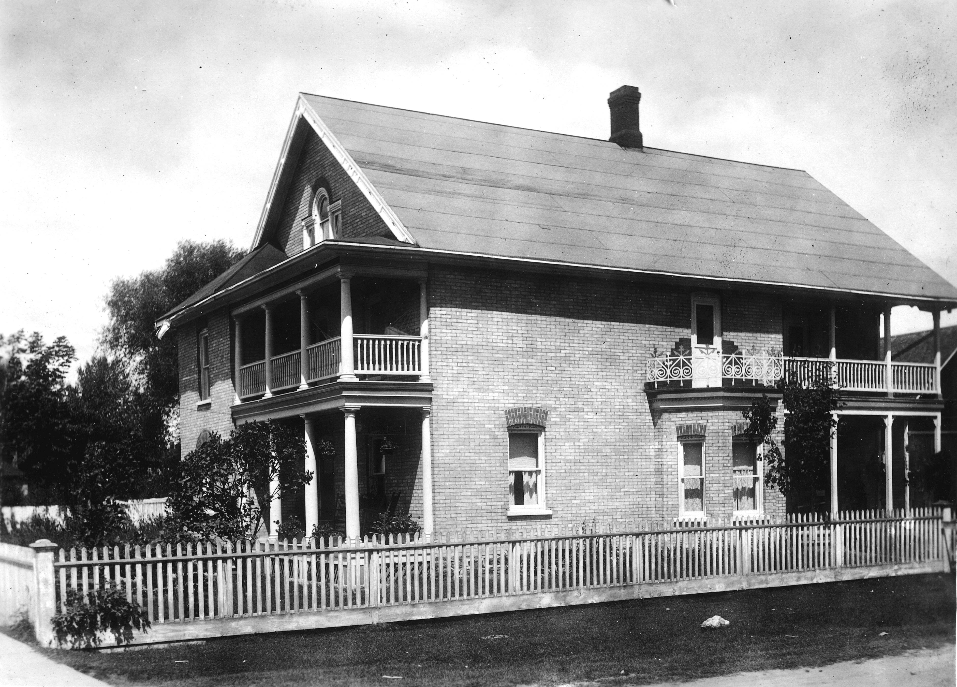 Two story brick house with covered porches at the front and side of the house on both the upper and lower levels. Picket fence surrounding the property.