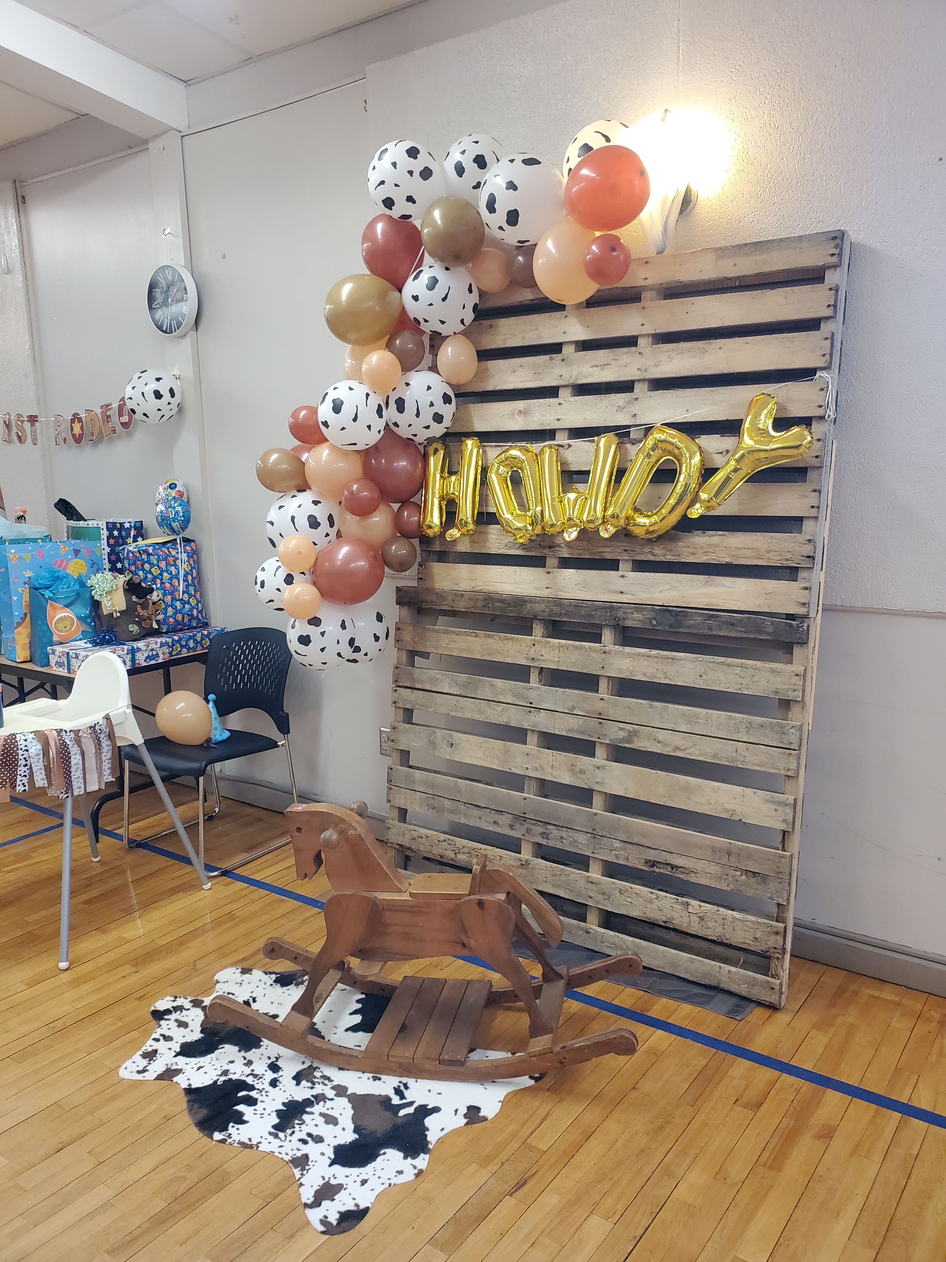 Wooden pallets leaned up against a wall with gold letters and the word Howdy across. Balloons, rocking horse and cow hide set up for child's birthday party.
