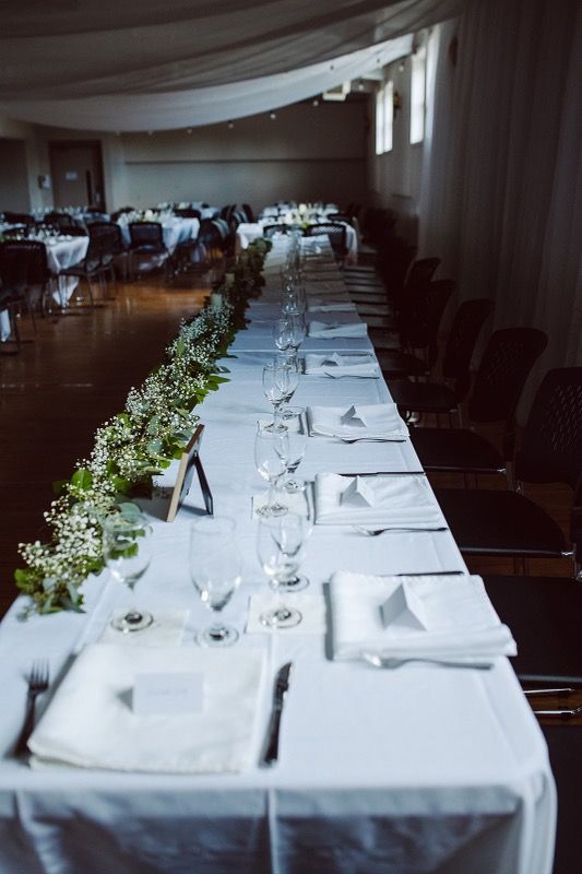 Long white table set up for a wedding reception. White linens and table cloths, greenery on one side.